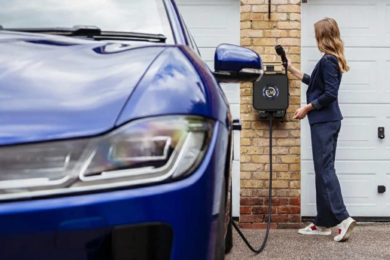 Donna che collega il cavo di ricarica di una wallbox domestica installata sul muro davanti al garage, con un'auto elettrica in primo piano